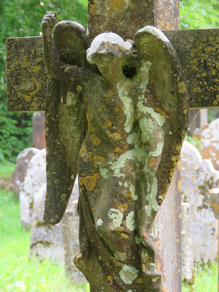 A lichen covered and eroded stone angel affixed to a stone cross headstone.